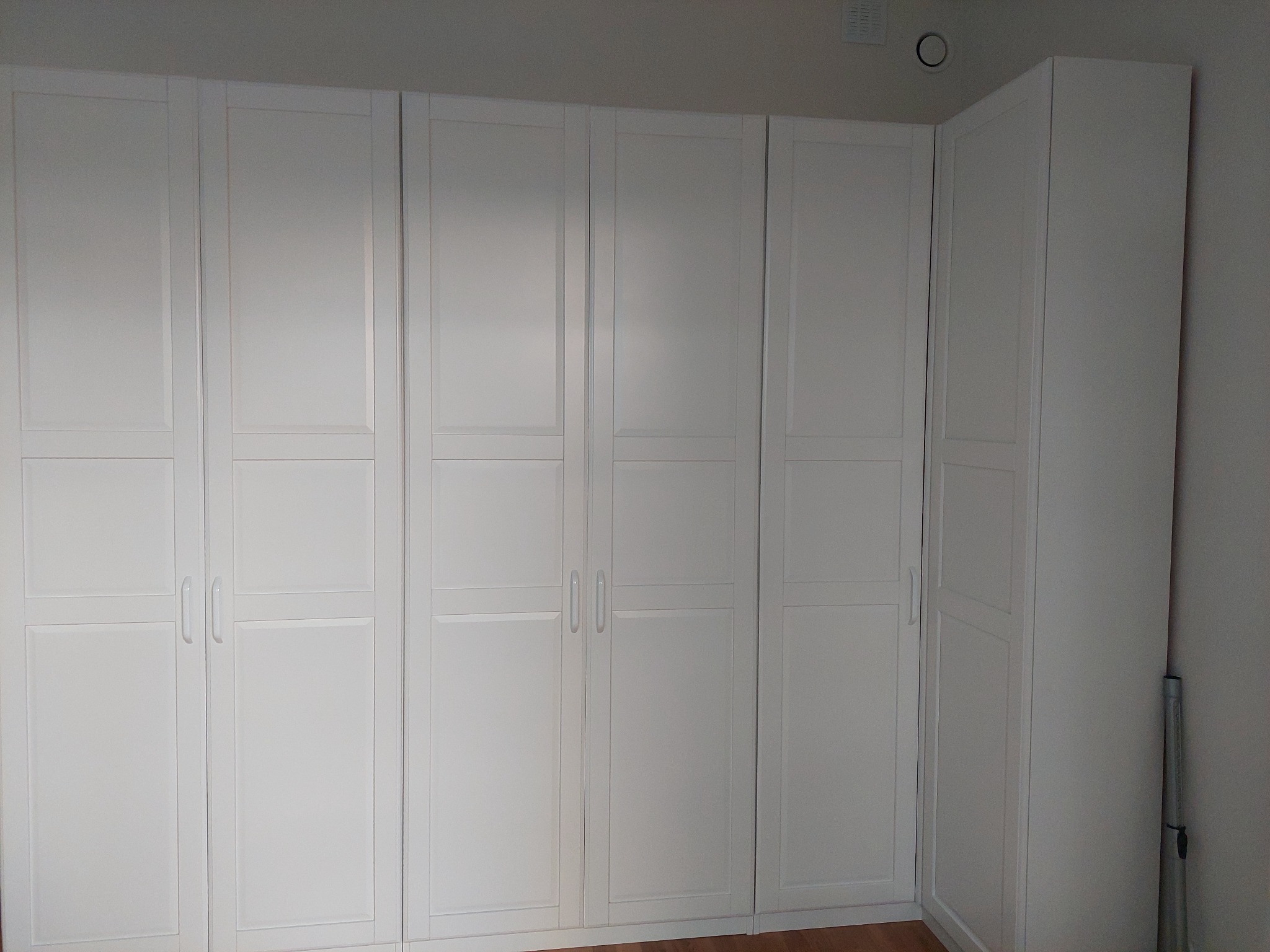 Biała zabudowa szaf wnękowych z panelowymi drzwiami, widoczny fragment podłogi i rura przy ścianie.