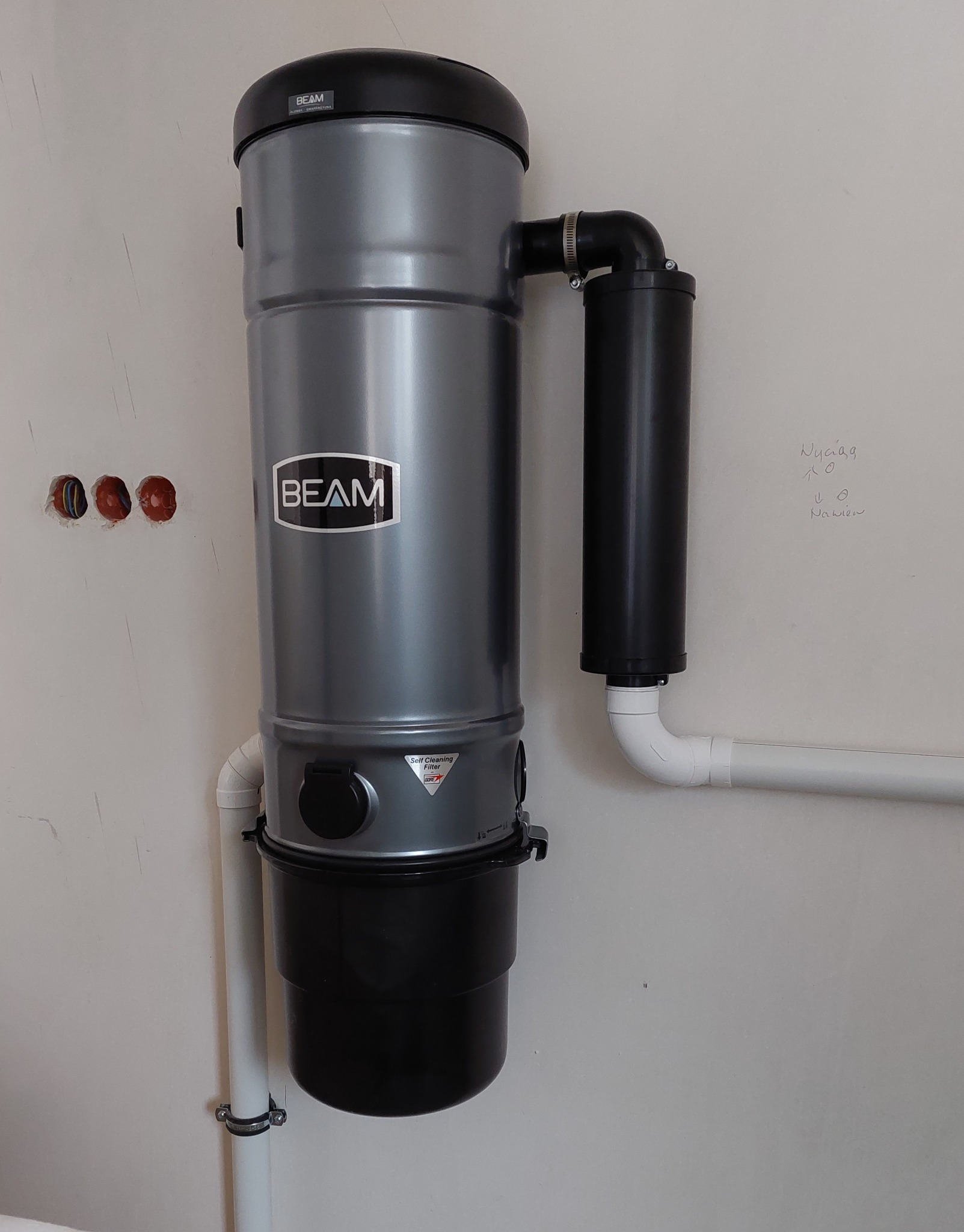 Szary odkurzacz centralny marki BEAM zamontowany na białej ścianie, widoczne rury instalacyjne i niedokończone otwory elektryczne w ścianie.