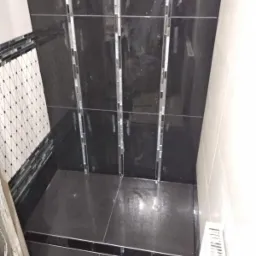 Wykończony brodzik z czarnymi płytkami, dekoracyjnymi listwami mozaikowymi i odpływem liniowym, widok z góry, w tle fragment ściany z biało-czarną mozaiką.