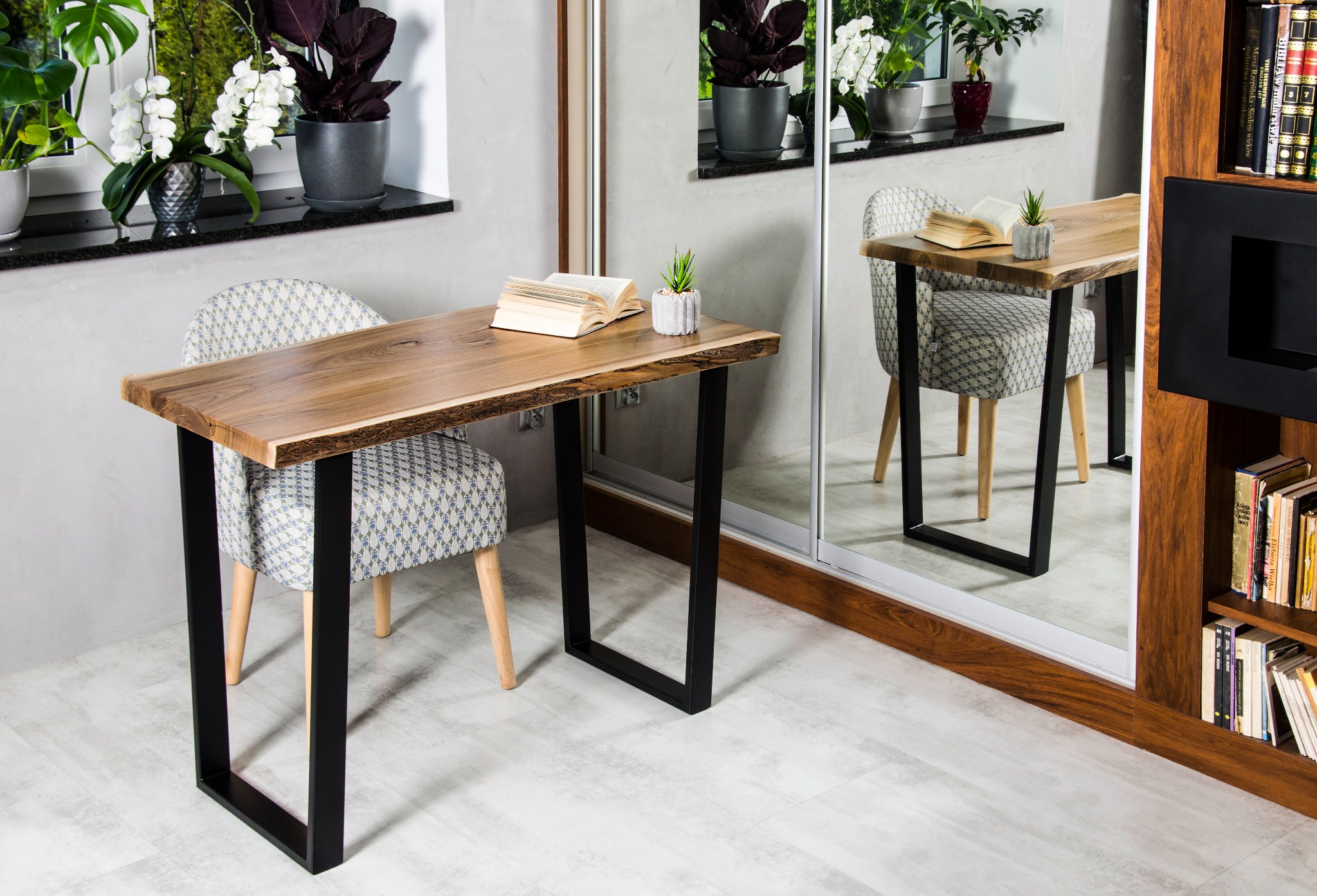 Biurko z blatem z litego drewna o nieregularnych krawędziach, czarne metalowe nogi, krzesło z tapicerką w geometryczny wzór, otwarta książka i sukulent w betonowej doniczce na blacie, w tle okno...