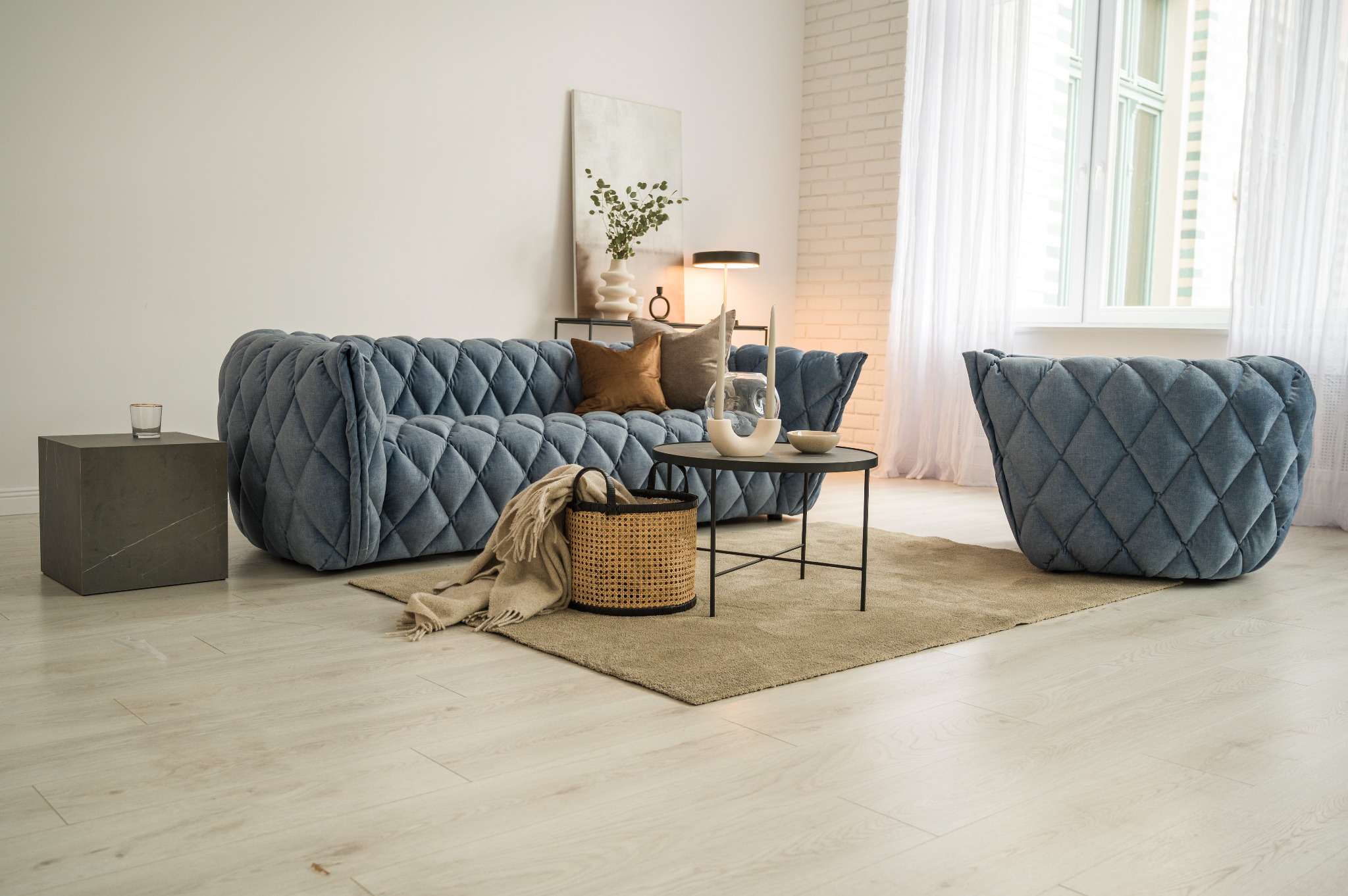 Jasne wnętrze salonu z pikowaną sofą i fotelem w odcieniach niebieskiego, stolikiem kawowym z dekoracjami, koszem z pledem oraz ceglaną ścianą w tle.