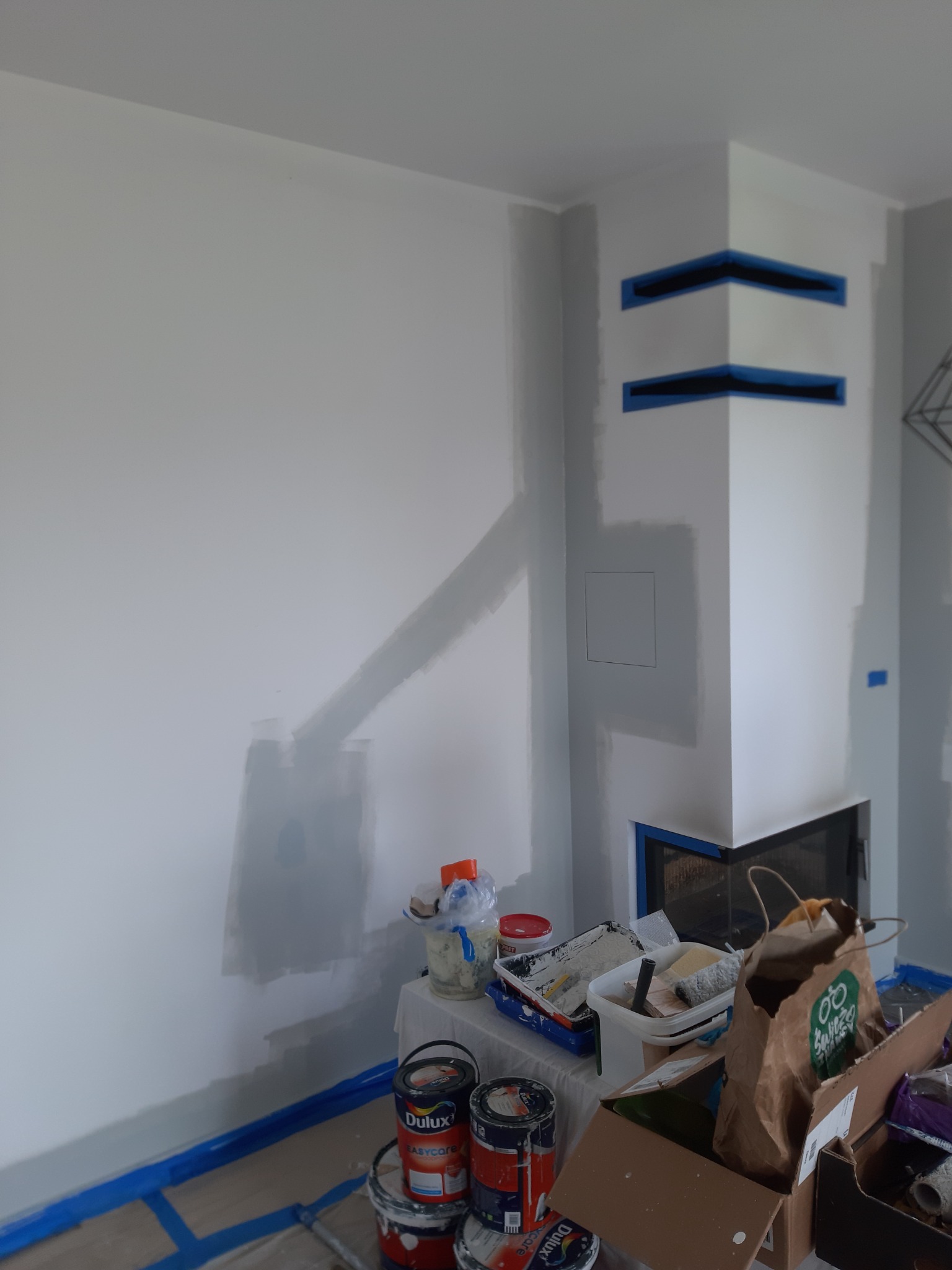 Świeżo pomalowana szara ściana z fragmentami zabezpieczonymi taśmą malarską, widoczne puszki z farbą Dulux i narzędzia malarskie na stole, przygotowane do dalszych prac w pomieszczeniu z kominkiem.