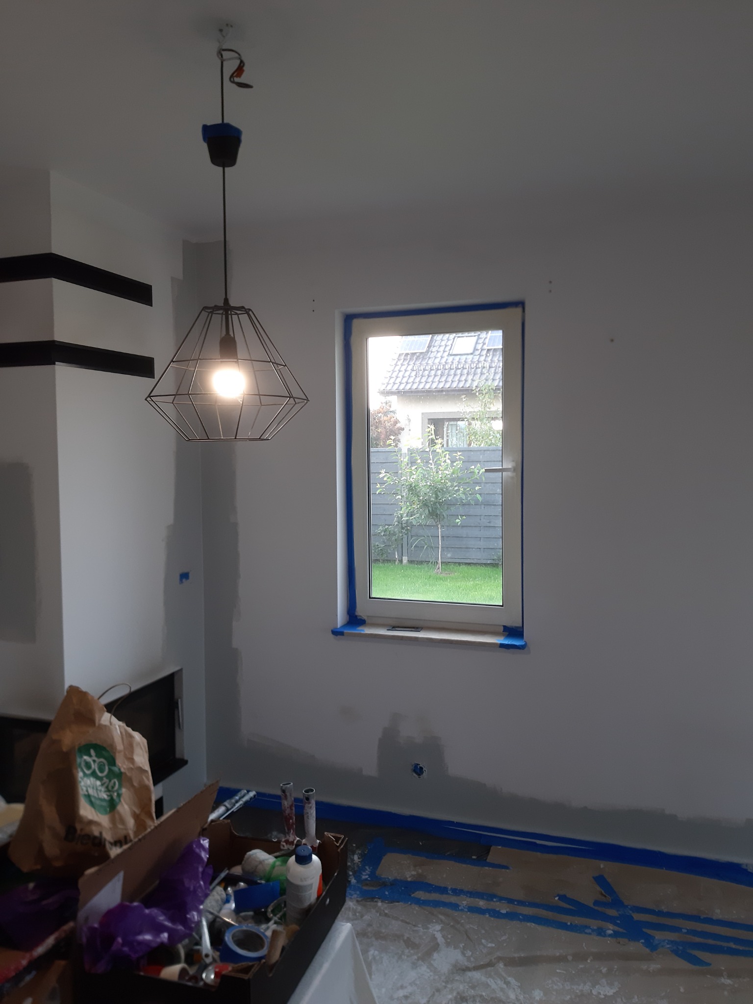 Pokój w trakcie malowania: ściany częściowo pokryte farbą, okno zabezpieczone taśmą malarską, podłoga zakryta folią ochronną, widoczna lampa wisząca i narzędzia malarskie.