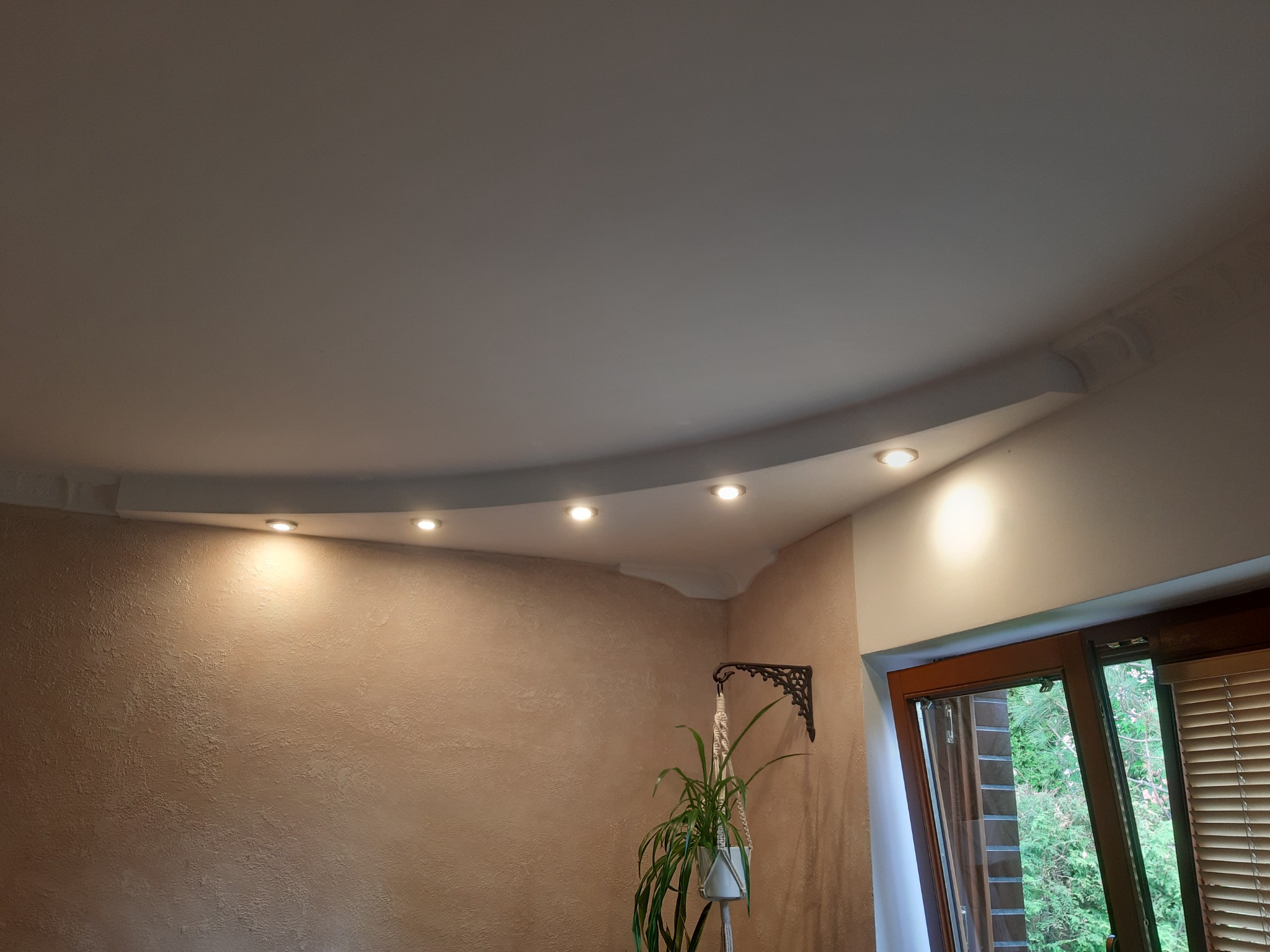 Kremowa ściana z dekoracyjną fakturą, podświetlana listwa sufitowa z wbudowanymi punktowymi lampami, widoczny fragment okna z żaluzjami i doniczkowa roślina wisząca na ozdobnym wieszaku.