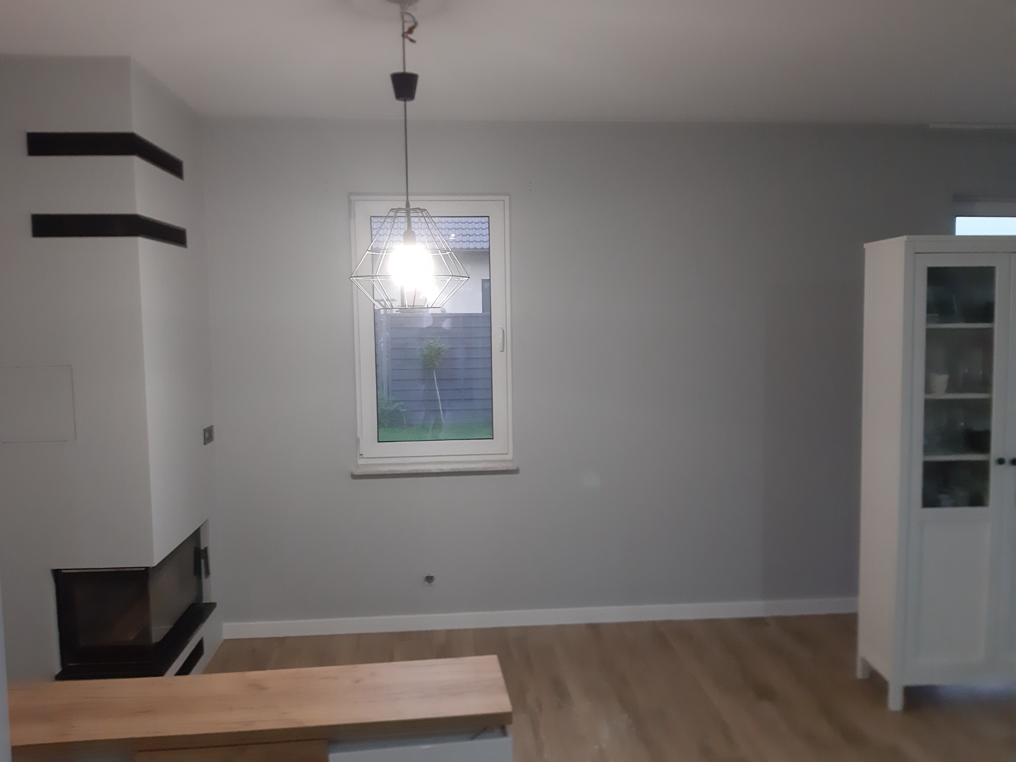 Jasnoszare ściany w pokoju po świeżym pomalowaniu, widoczny nowoczesny kominek z czarnymi elementami, drewniana podłoga, okno z widokiem na zewnątrz i wisząca lampa w geometrycznym stylu, biała...