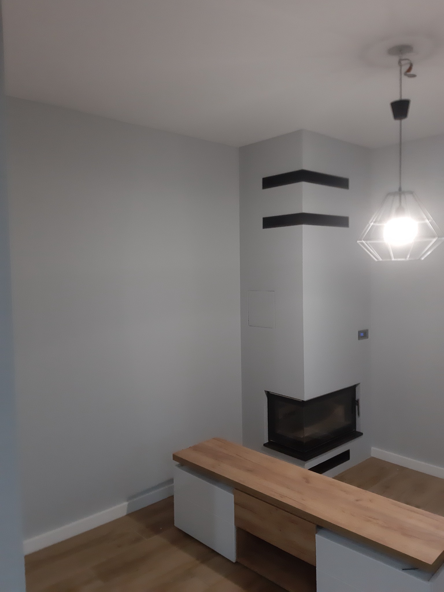 Jasnoszara ściana z wbudowanym kominkiem narożnym z czarnymi akcentami, drewniany blat, nowoczesna lampa wisząca w pomieszczeniu po odświeżeniu.