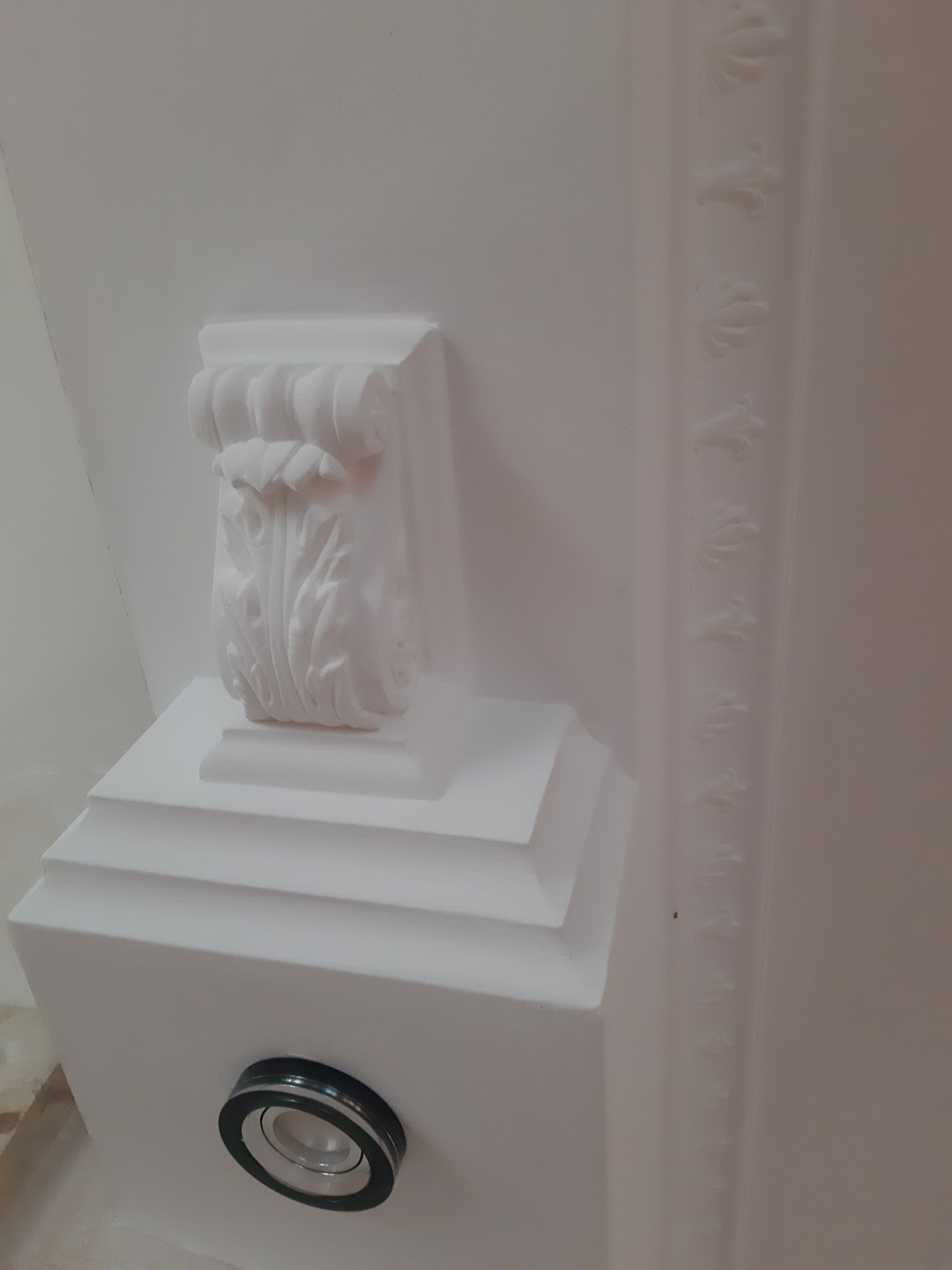 Biały, ozdobny wspornik sufitowy z roślinnym ornamentem, umieszczony nad wbudowanym punktem oświetleniowym w suficie, obok dekoracyjnej listwy sufitowej z delikatnym wzorem.