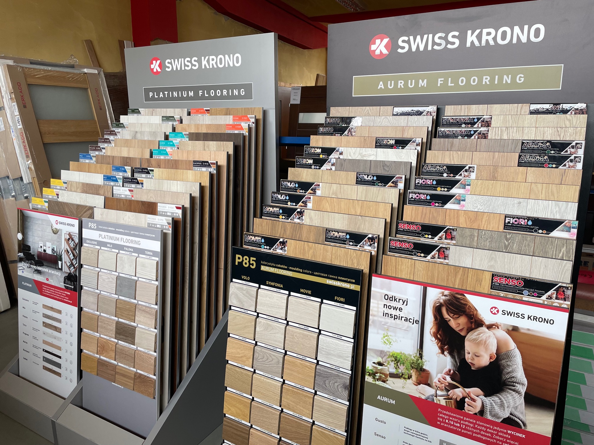 Ekspozycja paneli podłogowych Swiss Krono z serii Platinium i Aurum, prezentacja różnych wzorów i kolorów, widoczne próbki z etykietami i zdjęciem aranżacji z dzieckiem.