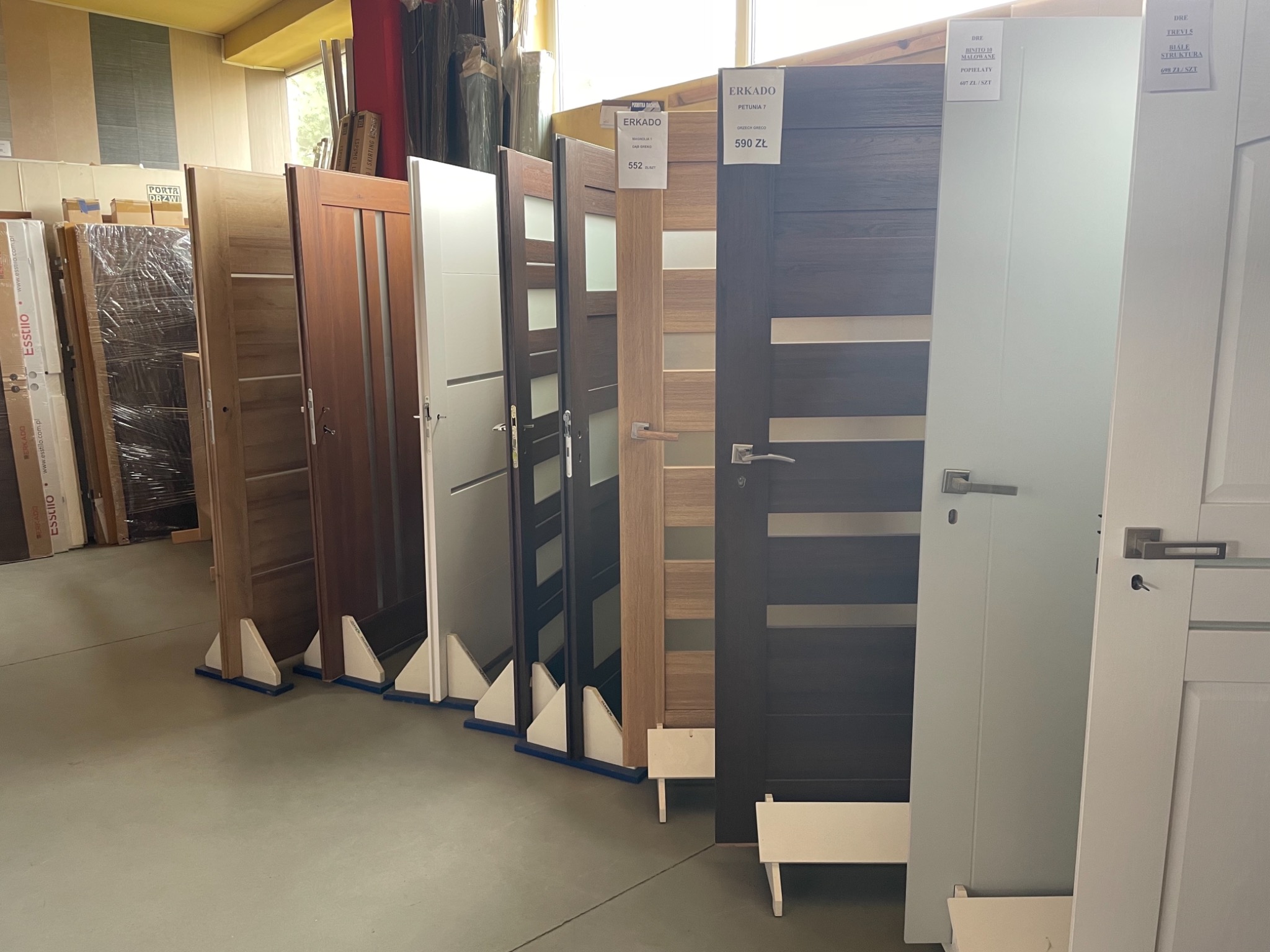 Ekspozycja różnych modeli drzwi wewnętrznych w sklepie, widoczne drzwi z przeszkleniami, w odcieniach brązu, szarości i bieli, z cenami i nazwami modeli.