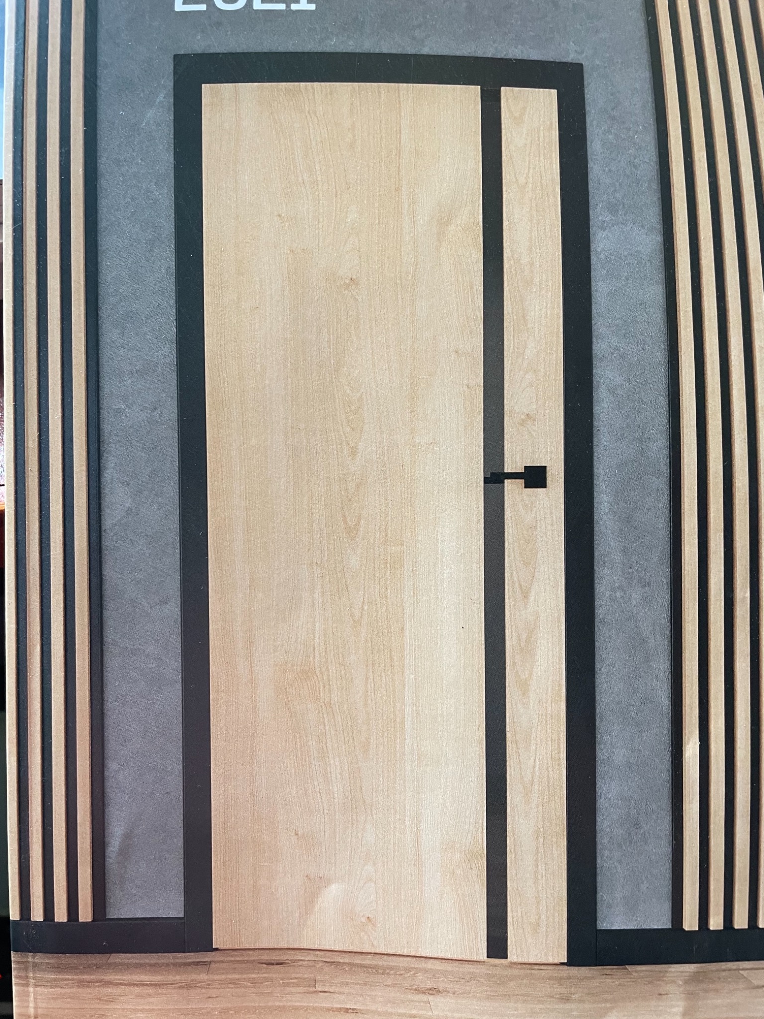 Nowoczesne drzwi wewnętrzne z jasnego drewna, z czarnymi wstawkami i minimalistyczną klamką na tle szarej ściany z drewnianymi lamelami po bokach.