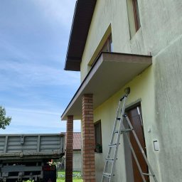 JKR - Żółta elewacja domu z ceglanymi kolumnami ganku, drabina oparta o ścianę, obok leży pies. W tle widoczny samochód ciężarowy i fragment budynku.