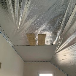 JKR - Skośne poddasze z izolacją termiczną, folią aluminiową i płytami gipsowo-kartonowymi. Widoczne drewniane elementy konstrukcyjne i okno.