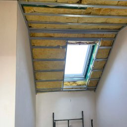 JKR - Skośny sufit poddasza z oknem dachowym w trakcie ocieplania wełną mineralną, widoczne metalowe profile i rusztowanie. Jasne wnętrze w trakcie prac wykończeniowych.