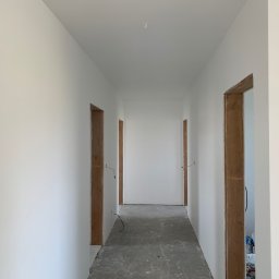 JKR - Biały korytarz po remoncie z surowymi ościeżnicami drzwi i wystającymi przewodami elektrycznymi. Świeżo pomalowane ściany i surowa podłoga.