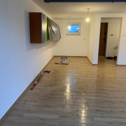 JKR - Jasne wnętrze po remoncie: świeżo pomalowane ściany, drewnopodobna podłoga z płytek, poziomica przy ścianie, puszka farby. Widoczne zabezpieczone meble.