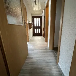 Długi korytarz z panelami podłogowymi w odcieniach szarości, drewniane drzwi z przeszkleniami na końcu, biała boazeria na suficie, oraz widoczne ościeżnice drzwiowe w jasnym drewnie.