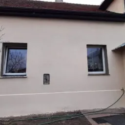 Elewacja domu jednorodzinnego po nałożeniu jasnobeżowej gładzi, widoczne dwa okna z białymi ramami, mała wnęka w ścianie, obok budynku rośnie drzewo bez liści, na dole wąż ogrodowy.