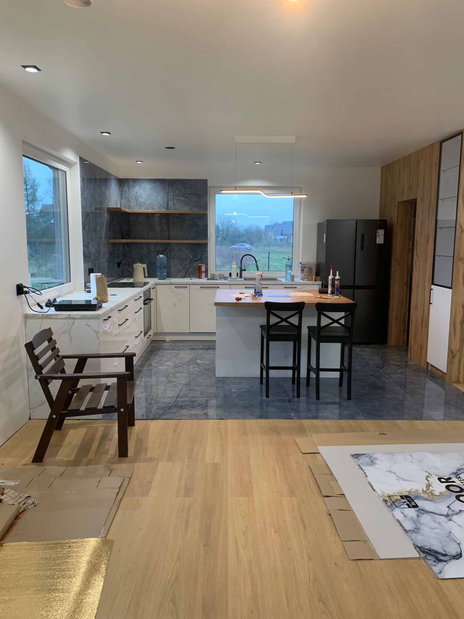 Nowoczesna kuchnia w trakcie wykończenia: białe szafki, marmurowe blaty, drewniane półki i podłoga. Widoczne materiały wykończeniowe i krzesło.