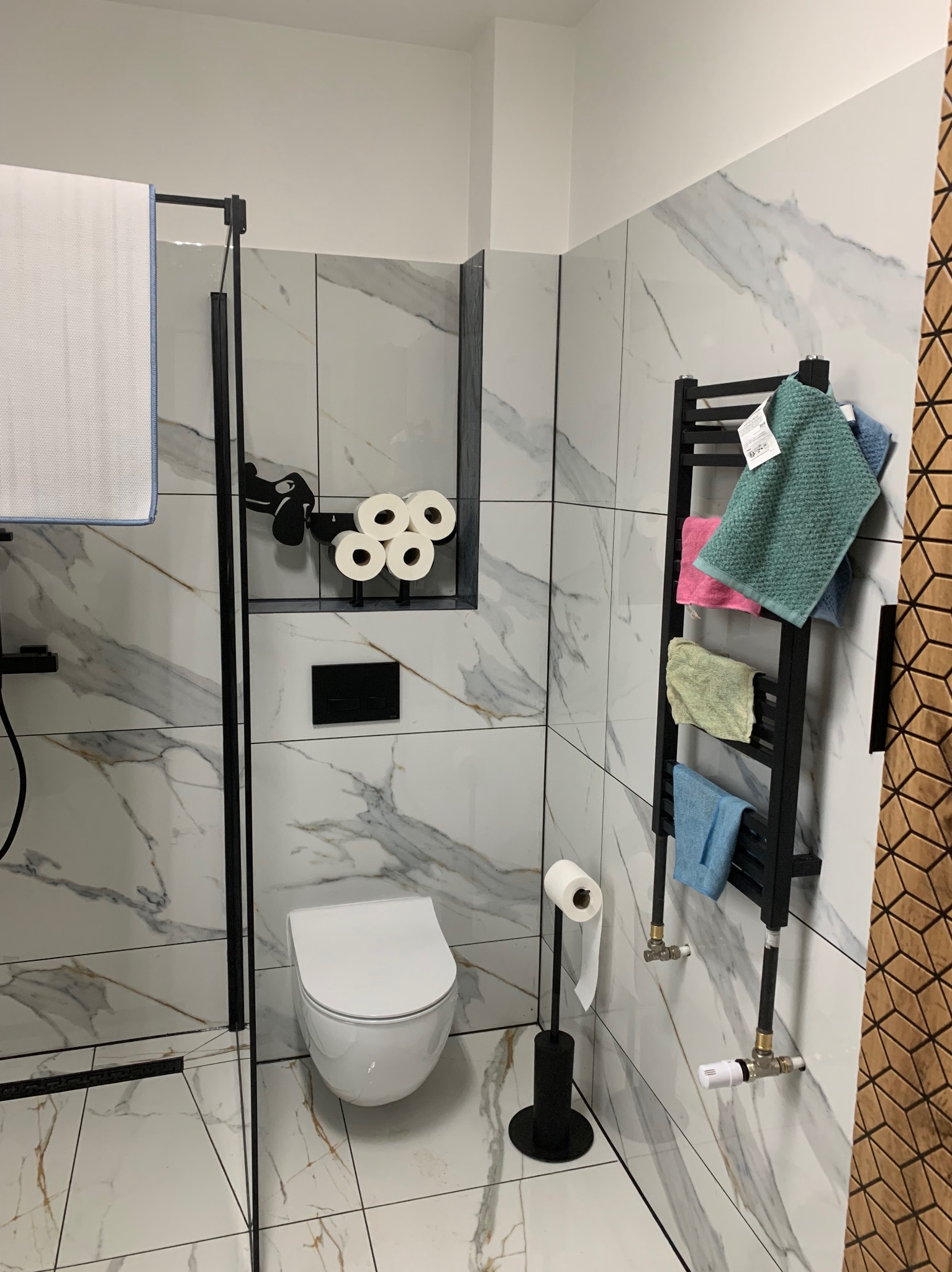 Nowoczesna łazienka z marmurowymi płytkami, czarnymi akcentami i grzejnikiem drabinkowym z ręcznikami. Minimalistyczny design z wiszącą toaletą i dekoracyjnym uchwytem na papier.