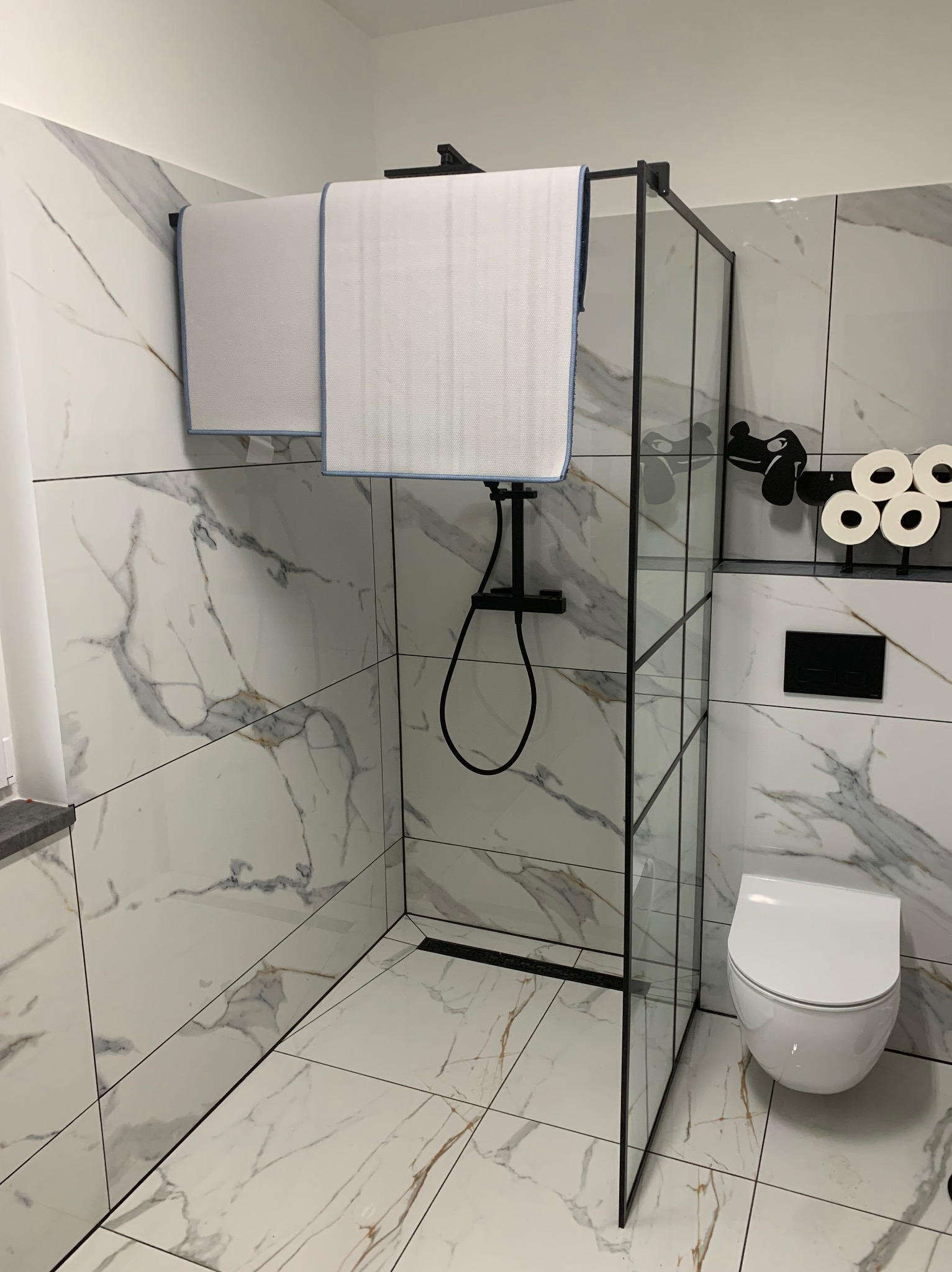 Nowoczesna łazienka z kabiną prysznicową typu walk-in, białe płytki imitujące marmur z delikatnymi złotymi żyłkami, czarna armatura i akcesoria.