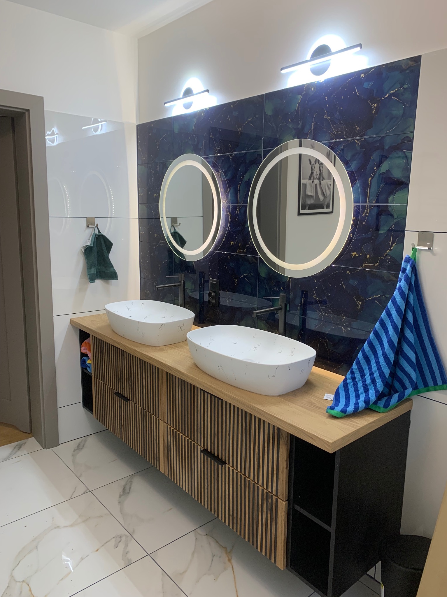 Nowoczesna łazienka z dwoma umywalkami nablatowymi, podświetlanymi lustrami i płytkami imitującymi marmur. Drewniana szafka pod umywalkami z czarnymi akcentami.