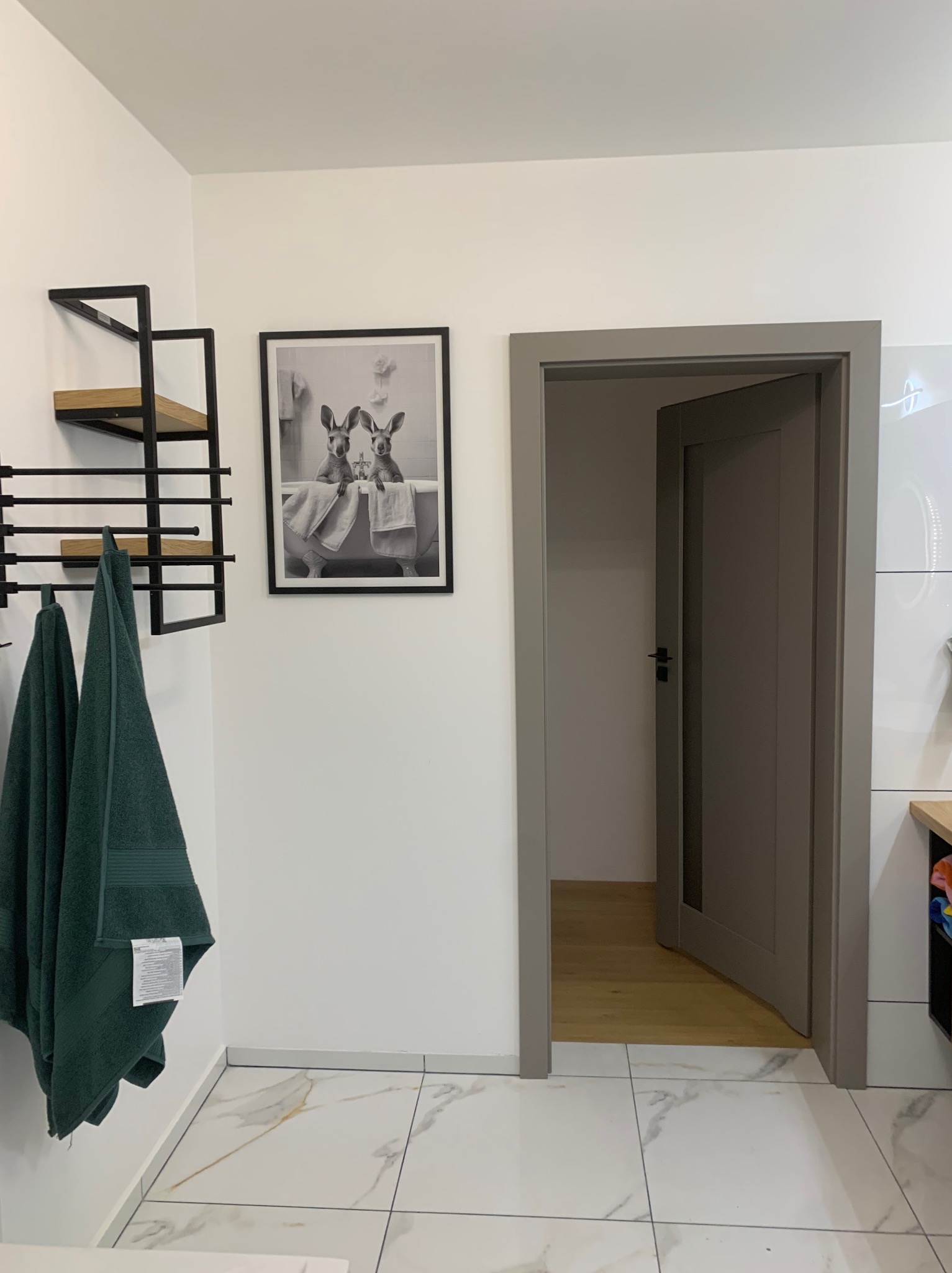 Nowoczesna łazienka z marmurową podłogą, szarą ramą drzwi i ręcznikami w kolorze butelkowej zieleni. Na ścianie grafika w czarnej ramie z motywem zwierzęcym.