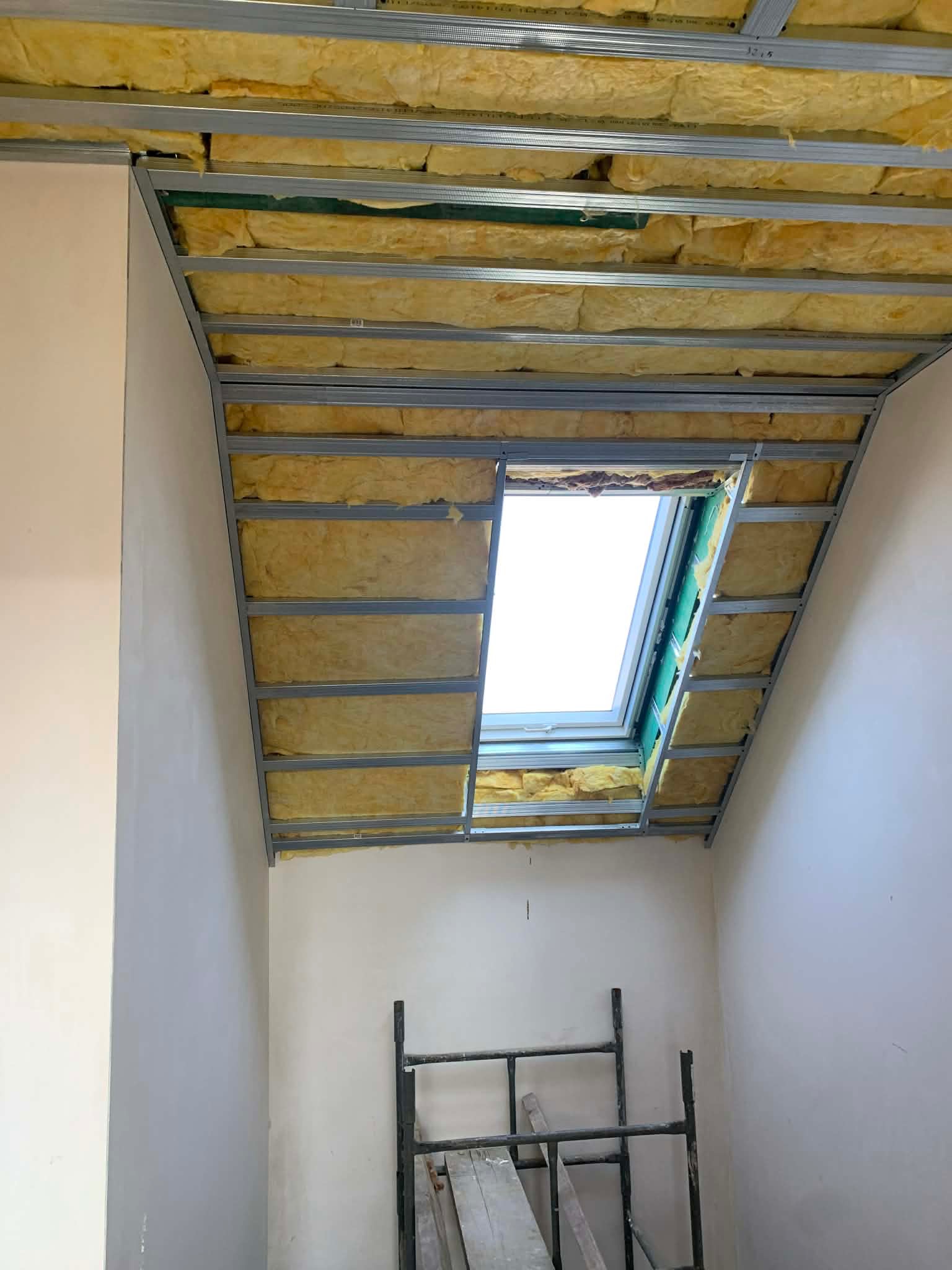 Skośny sufit poddasza z oknem dachowym w trakcie izolacji wełną mineralną, widoczna metalowa konstrukcja i fragment rusztowania.