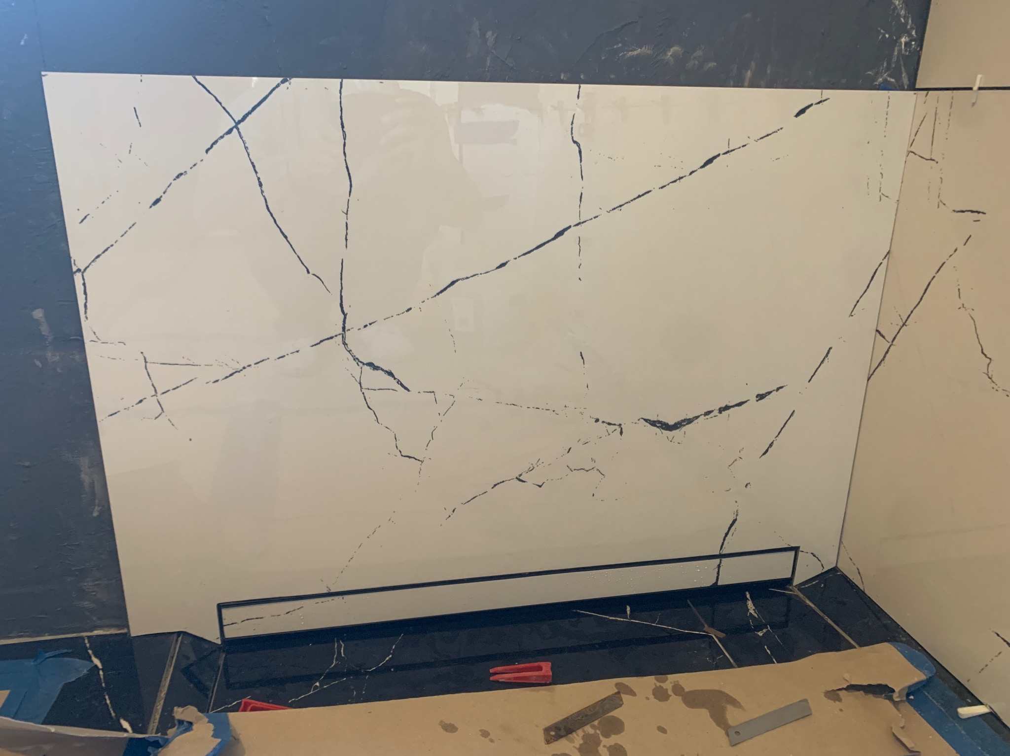 Montaż wielkoformatowej płyty imitującej marmur na ścianie w łazience. Widoczny odpływ liniowy i narzędzia glazurnicze. Prace wykończeniowe w toku.