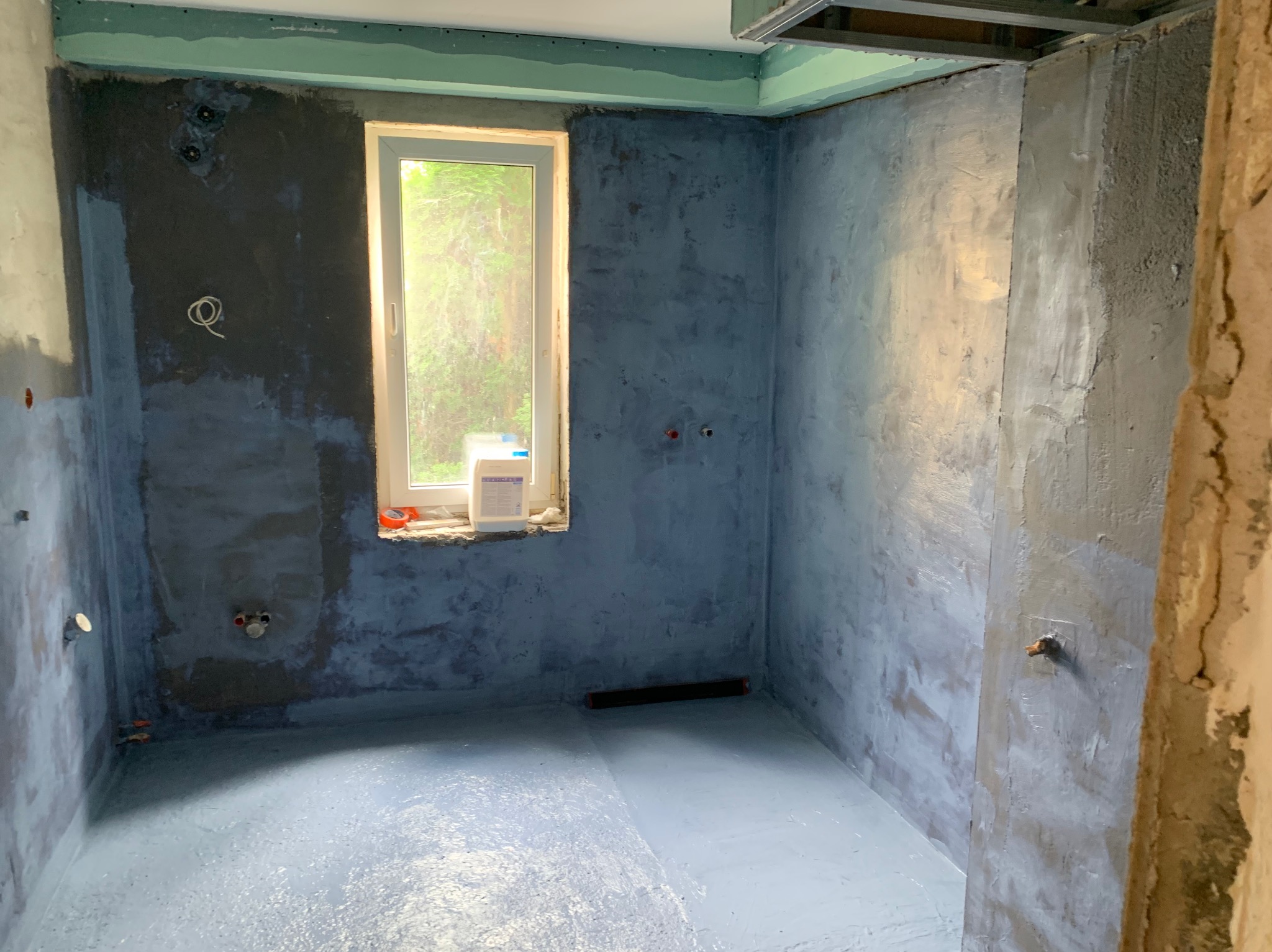 Hydroizolacja w łazience w trakcie prac. Widoczne zabezpieczone ściany i podłoga, przygotowane pod dalsze etapy wykończenia. Okno z widokiem na zieleń.