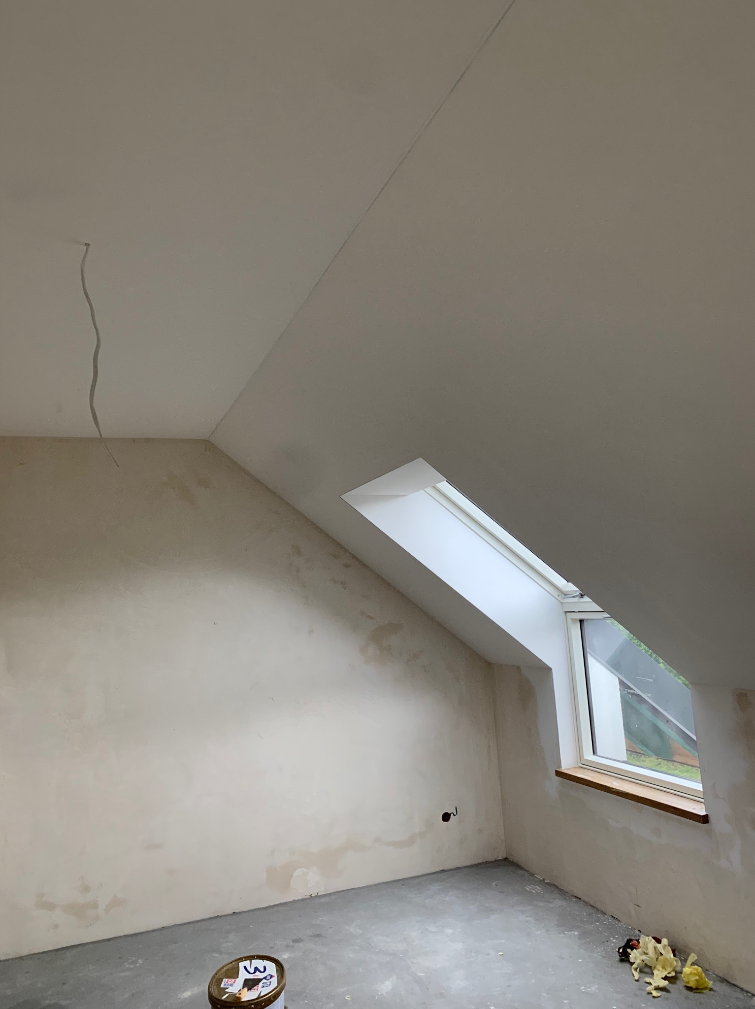 Poddasze ze skośnym oknem dachowym w trakcie prac wykończeniowych. Ściany i sufit po nałożeniu gładzi, widoczne niedoskonałości i przygotowanie do malowania.