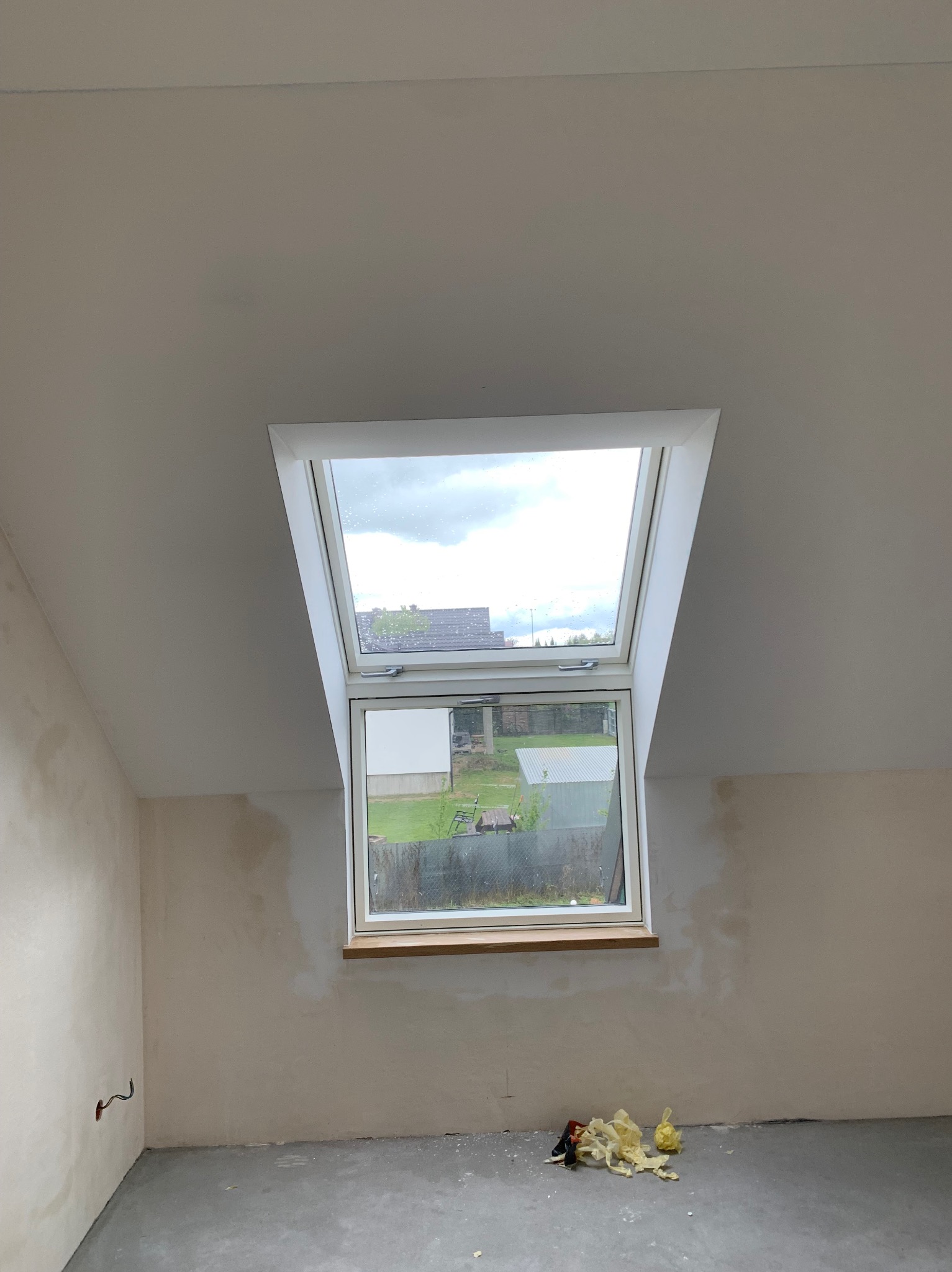 Poddasze po gipsowaniu ścian, z oknem dachowym i widokiem na zewnątrz. Ściany przygotowane do malowania, widoczne ślady po łatach gipsowych. Na podłodze resztki materiałów.