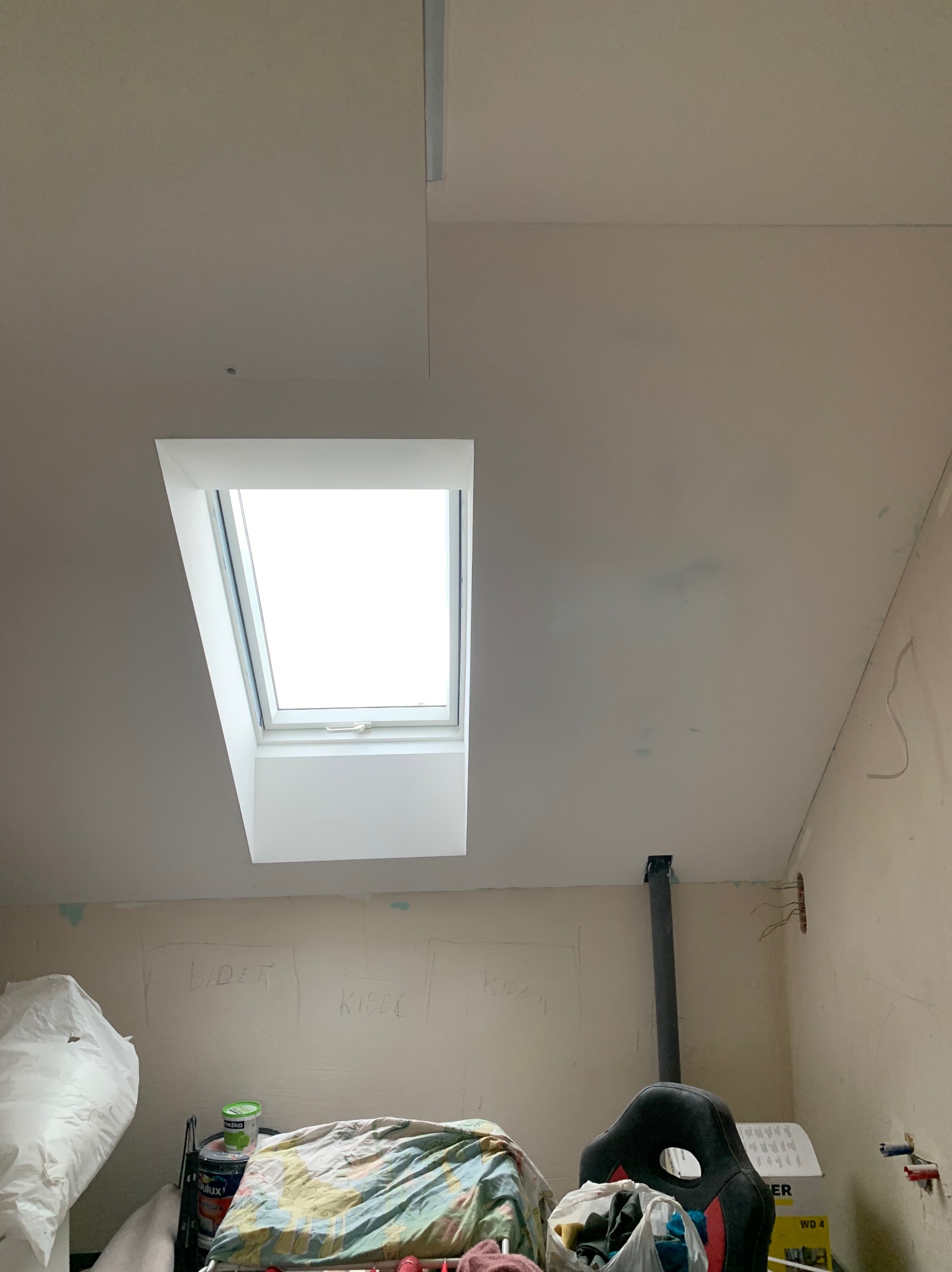 Poddasze z oknem dachowym po montażu płyty gipsowej na suficie, widoczne ślady po szpachlowaniu i przygotowaniu do malowania. Na ścianach widoczne napisy.