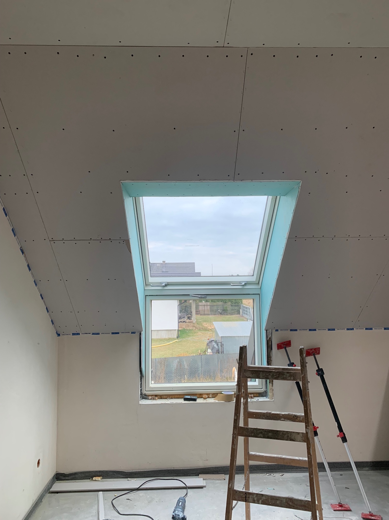 Poddasze z oknem dachowym w trakcie montażu płyt gipsowo-kartonowych. Widoczna drabina, narzędzia i częściowo wykończone ściany. Charakterystyczne niebieskie zabezpieczenia.