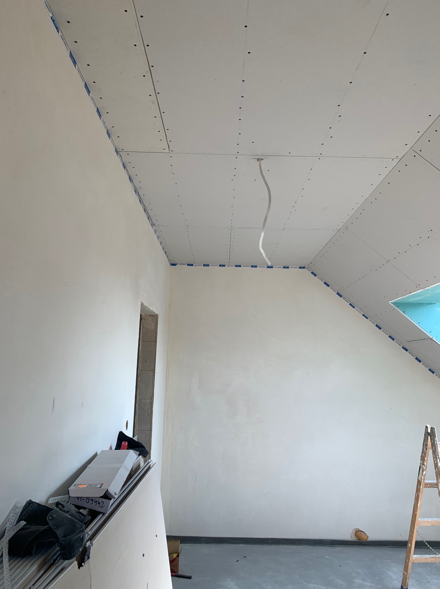 Poddasze w trakcie prac: ściany po gładziowaniu, zamontowane płyty karton-gips na skosach sufitu, widoczne łączenia płyt. Na podłodze narzędzia i drabina.