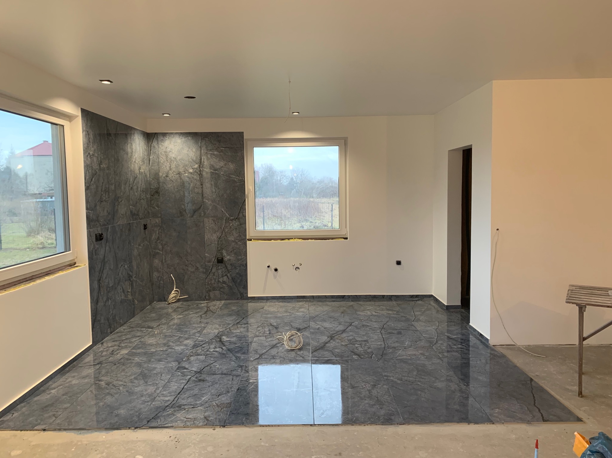 Wykończone wnętrze z szarymi płytkami imitującymi marmur na podłodze i ścianie, widoczne okna i przygotowane punkty elektryczne.