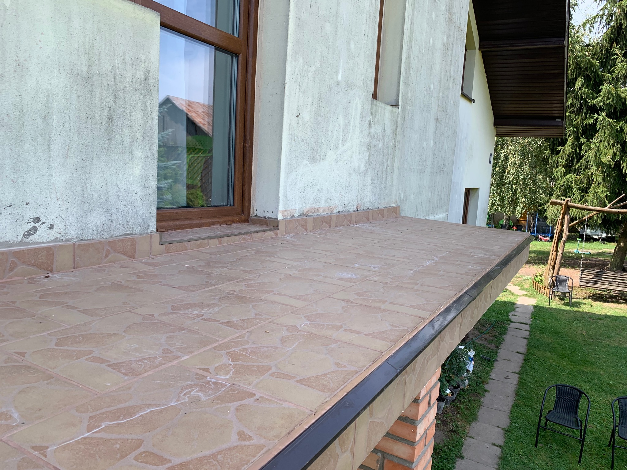 Wykończony taras z brązowymi płytkami ceramicznymi, widok z góry. Na ścianie widoczne okno z brązową ramą. W tle zieleń ogrodu i huśtawka.