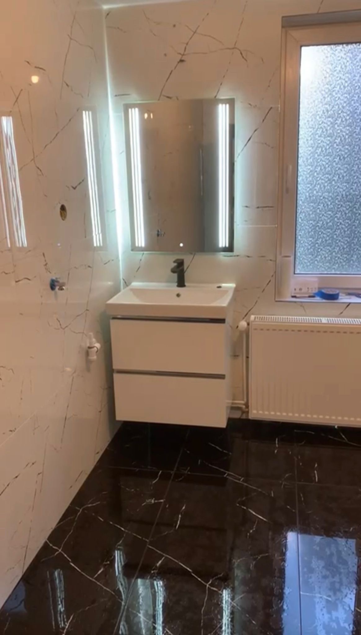 Wykończona łazienka z czarno-białymi marmurkowymi płytkami, białą szafką z umywalką i podświetlanym lustrem. Nowoczesny grzejnik pod oknem.