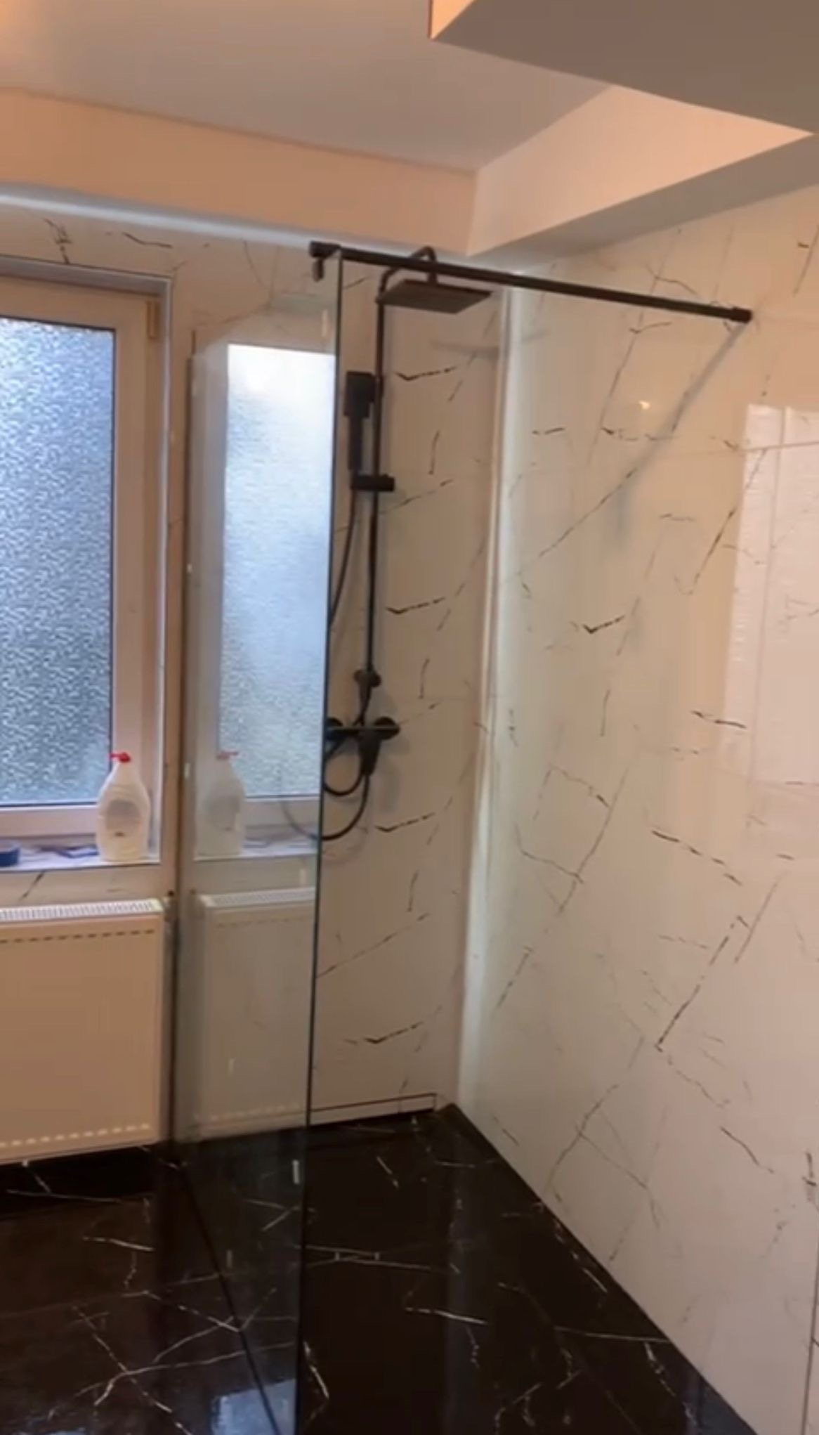 Wykończona łazienka w Gozdawie: kabina prysznicowa typu walk-in z czarną armaturą, marmurowe płytki na ścianach i podłodze, okno z parapetem.