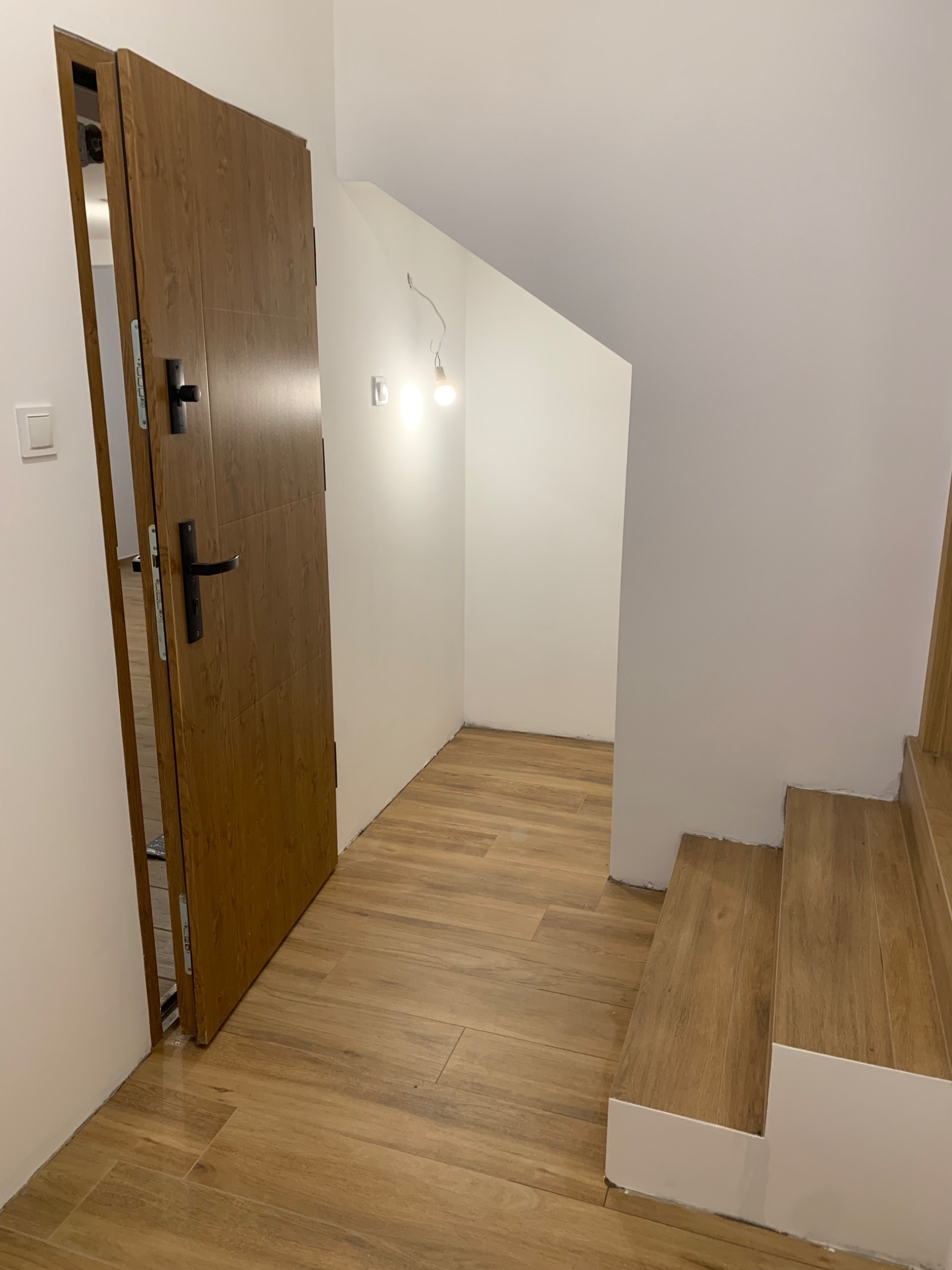 Otwarta drewniana okleina drzwiowa, jasne płytki podłogowe imitujące drewno, białe ściany i schody. Minimalistyczne wnętrze po remoncie.