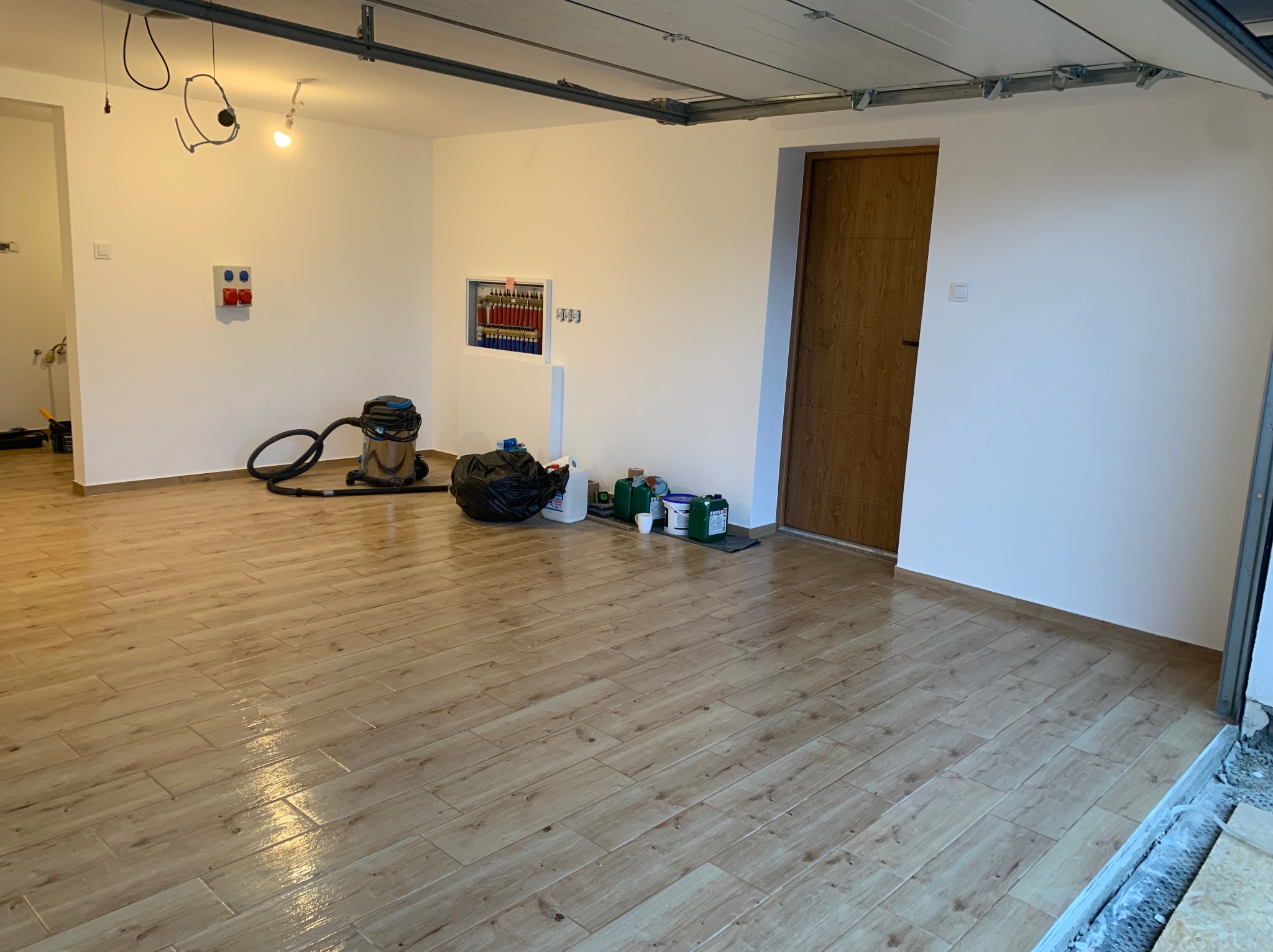 Wykończone wnętrze garażu z jasnymi płytkami imitującymi drewno, świeżo pomalowanymi ścianami i instalacją elektryczną. Widoczny odkurzacz i pojemniki z farbą.