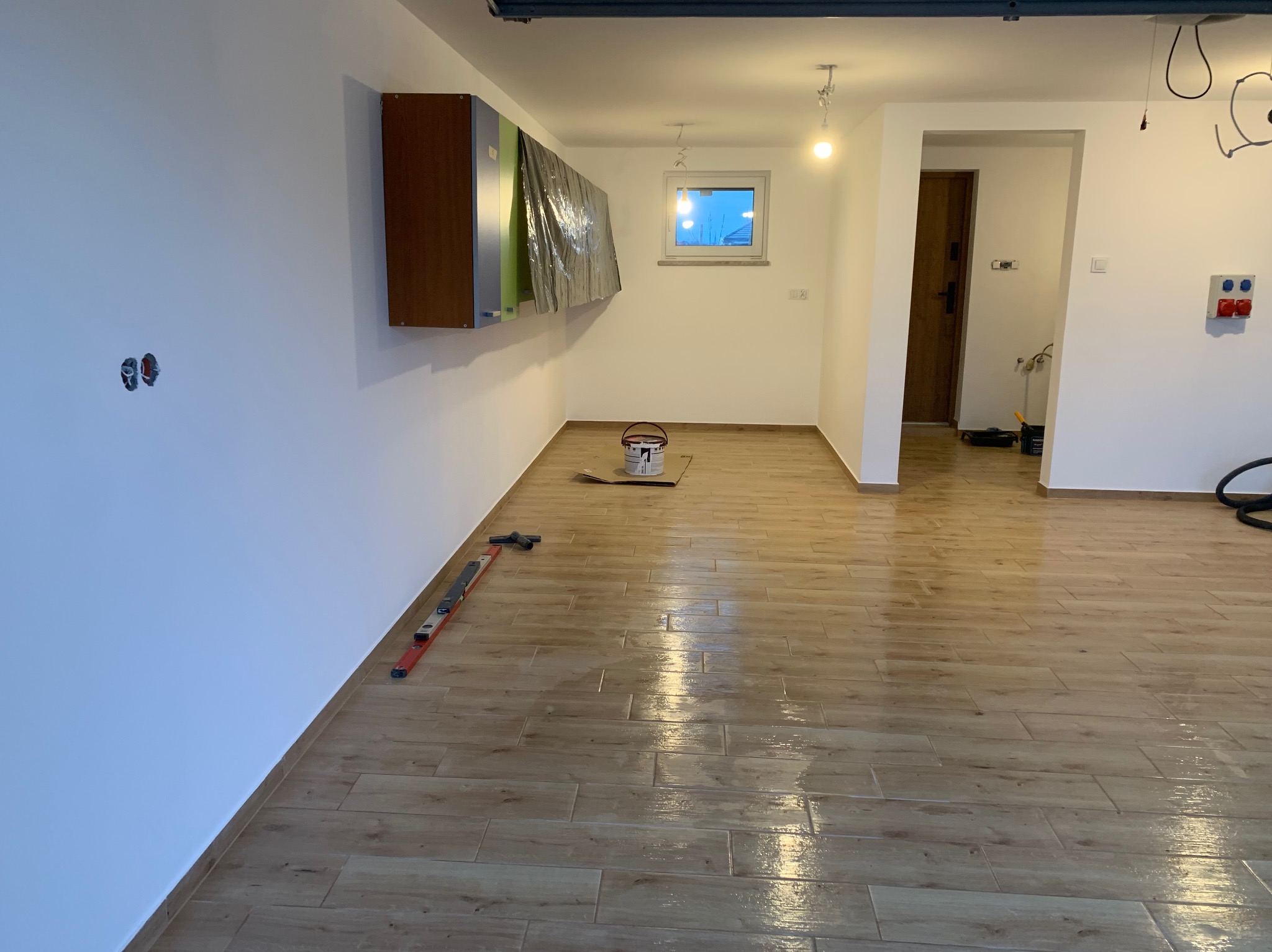 Jasne wnętrze po remoncie: świeżo pomalowane ściany, drewnopodobna podłoga z płytek, poziomica przy ścianie, puszka farby. Widoczne zabezpieczone meble.