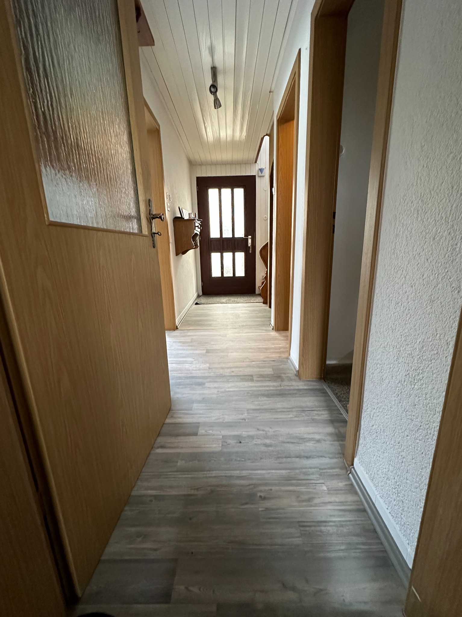 Długi korytarz z panelami podłogowymi w odcieniach szarości, drewniane drzwi z przeszkleniami na końcu, biała boazeria na suficie, oraz widoczne ościeżnice drzwiowe w jasnym drewnie.