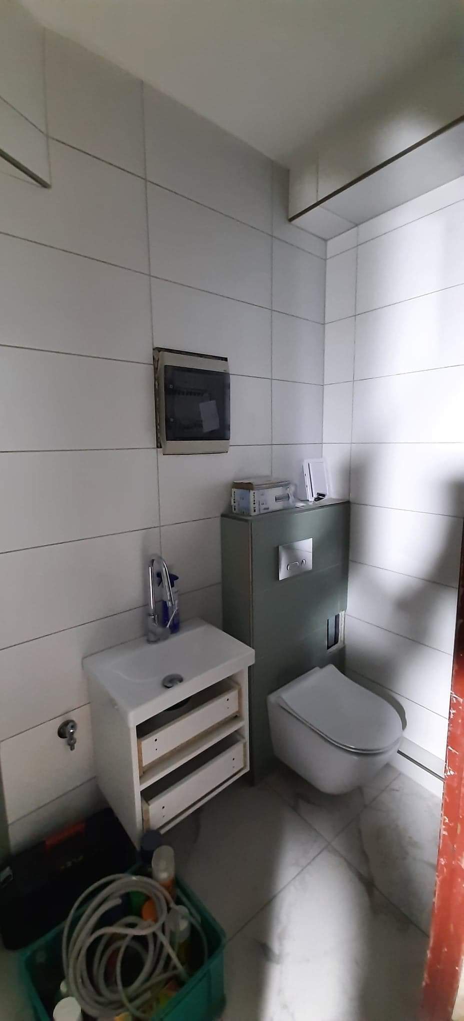 Biała łazienka w trakcie remontu: widoczna toaleta podwieszana, umywalka z szafką bez frontów szuflad, skrzynka elektryczna na ścianie i pojemnik z narzędziami na podłodze.
