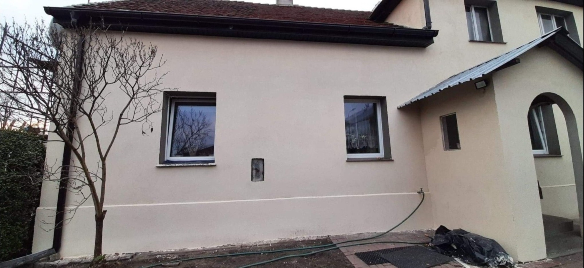 Elewacja domu jednorodzinnego po nałożeniu jasnobeżowej gładzi, widoczne dwa okna z białymi ramami, mała wnęka w ścianie, obok budynku rośnie drzewo bez liści, na dole wąż ogrodowy.