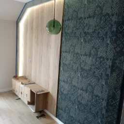 Wnętrze w trakcie remontu: fragment ściany z nałożoną tapetą w ciemnoniebieskim odcieniu z wzorem, obok drewnopodobna płyta z oświetleniem LED, zielona szklana lampa wisząca, częściowo widoczny...