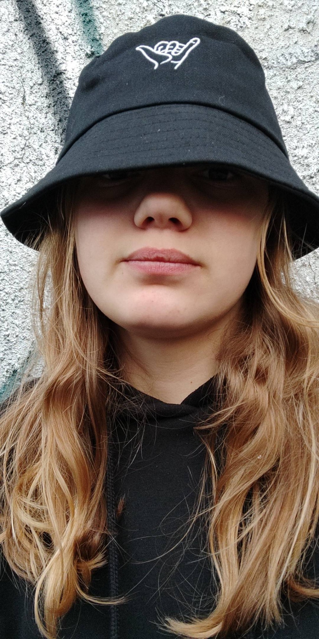 Portret kobiety w czarnym kapeluszu typu bucket z białym haftem, na tle szarej ściany z teksturą.