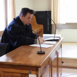 Mężczyzna w todze siedzi przy drewnianym stole w sali sądowej, czytając dokumenty pod mikrofonem.