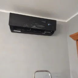 Czarny klimatyzator AUX zamontowany na szarej ścianie w pomieszczeniu, widoczny fragment drabiny i framuga drzwi.