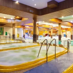 Wnętrze luksusowego spa z kilkoma owalnymi jacuzzi z żółtymi obudowami, metalowymi poręczami, mozaikowymi kolumnami, drewnianym sufitem i malowidłem ściennym z motywem wodospadu.