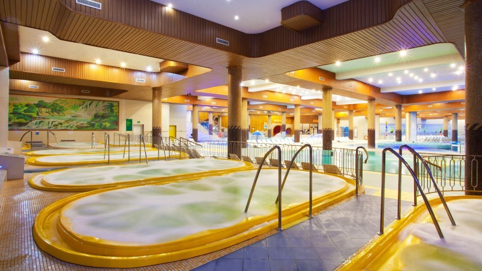 Wnętrze luksusowego spa z kilkoma owalnymi jacuzzi z żółtymi obudowami, metalowymi poręczami, mozaikowymi kolumnami, drewnianym sufitem i malowidłem ściennym z motywem wodospadu.